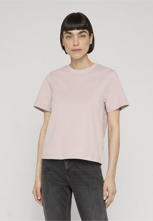 Femme aux cheveux courts et foncés portant un t-shirt rose clair à manches courtes et un jean noir sur un fond clair uni.