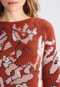 Strikket trøje med en rust-rød bundfarve og blomster i creme og sort. Den har en bred halsudskæring og blødt, tekstureret stof.