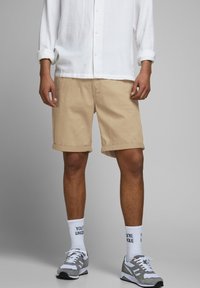 Pantalones cortos de algodón beige con dobladillos enrollados, combinados con zapatillas deportivas grises y calcetines blancos con texto negro. La camisa es de manga larga y blanca.