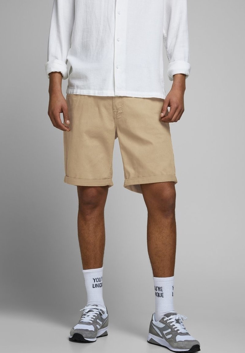 Pantalones cortos de algodón beige con dobladillos enrollados, combinados con zapatillas deportivas grises y calcetines blancos con texto negro. La camisa es de manga larga y blanca.