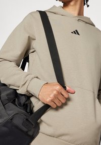 Felpa beige con logo nero sul petto, abbinata a una borsa a spalla nera con superficie texture e una tracolla.