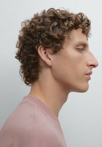 Vista profilo di una persona con capelli ricci castani, che indossa una camicia rosa chiaro con colletto arrotondato su uno sfondo neutro.