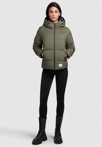 Olivengrøn pufferjakke med hætte, front lynlås og sorte accenter, parret med sorte leggings og snørebåndskampstøvler, mod en lys baggrund.