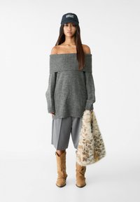 Grijze off-shoulder trui met geribbelde halslijn, grijze geplooide shorts, tan cowboylaarzen en een gestreepte nepbont tas. Marineblauw pet met tekst.