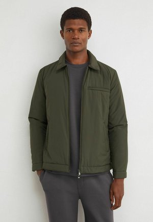 Junger Mann lehnt an einer weißen Wand, trägt eine dunkelgrüne Reißverschlussjacke über einem grauen Hemd und passenden grauen Hosen.