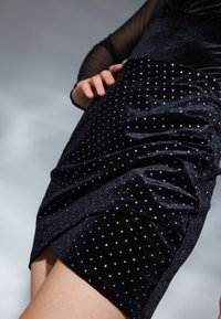 Robe en velours noir avec des pois argentés, présentant une silhouette ajustée et des détails froncés sur un côté. Les manches longues transparentes sont visibles.