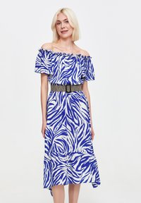 Kobieta z włosami w kolorze blond do ramion nosi niebieską i białą sukienkę w paski zebry z odkrytymi ramionami, podkreśloną czarnym plecionym paskiem.