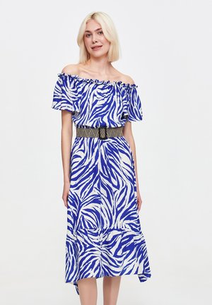 Kobieta z włosami w kolorze blond do ramion nosi niebieską i białą sukienkę w paski zebry z odkrytymi ramionami, podkreśloną czarnym plecionym paskiem.