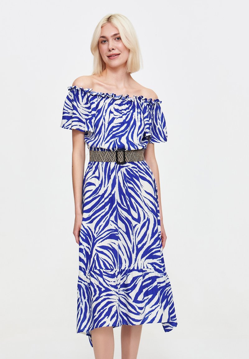 Kobieta z włosami w kolorze blond do ramion nosi niebieską i białą sukienkę w paski zebry z odkrytymi ramionami, podkreśloną czarnym plecionym paskiem.