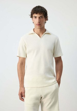 Hombre con una camiseta de punto texturizada crema de manga corta y pantalones cortos a juego, de pie frente a un fondo liso y claro.