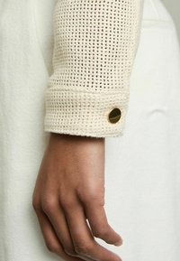 Crème-kleurige lange mouw kledingstuk met een gemosterd gebreid manchet, afgewerkt met een gouden knopaccent. Het lichaam is van glad materiaal.