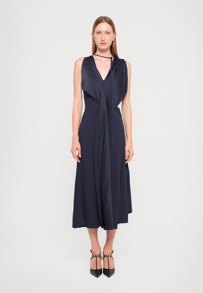 Robe en satin bleu marine avec un décolleté en V, sans manches ; présente un devant drapé et un détail de nouage autour du cou ; silhouette fluide, arrivée au genou.