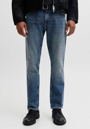 Homme portant un jean bleu délavé à jambes droites, une veste en cuir noire et des chaussures noires à lacets, debout devant un fond uni.