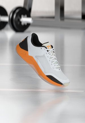 Weißer und orangener Sportschuh mit schwarzen Akzenten und Schnürsenkeln auf einem reflektierenden Fitnessboden, mit verschwommenen Hanteln im Hintergrund.