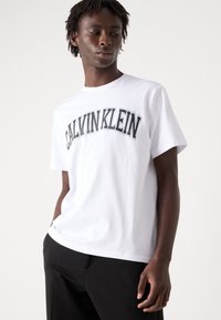 Hvit bomull t-skjorte med "CALVIN KLEIN" logo i svart og grått, rund hals, korte ermer og løs passform. Brukt sammen med svarte bukser.