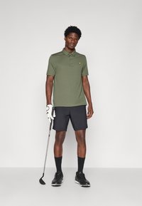Olīvu zaļš polo krekls ar īsu apkakli un dzeltenu logo, apvienots ar melnajām šortiem. Modelis tur golfu nūju, ģērbies melnās kurpēs un cimdiem.