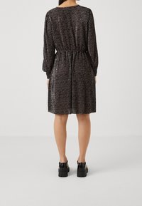 Kvinna står i en svart klänning med vita prickar, långa ärmar, åtsittande midja och svarta skor med tjock sula, vänd bort från kameran mot en enhetlig bakgrund.