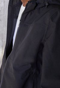 Černá voděodolná bunda s kapucí, zipovým zapínáním a kapsami. Vyrobena z texturované látky s viditelným švem a moderním designem.