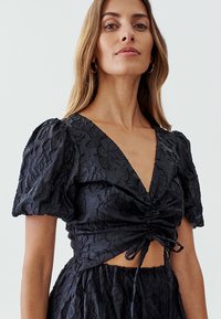 Czarny crop top w kwiatowy wzór z bufiastymi rękawami, dekoltem w serek oraz marszczeniem z detalem w postaci węzła z przodu. Teksturowana tkanina dodaje głębi.