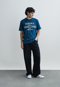 T-shirt de algodão azul com "Versace Milano" e design gráfico, combinada com calças largas pretas e ténis brancos, com um fundo neutro.