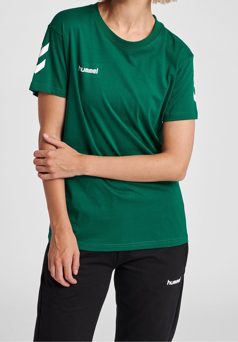 Camiseta de algodón verde con cuello redondo, mangas cortas y logo "hummel" en blanco. Presenta acentos en forma de chevrón en blanco en las mangas.