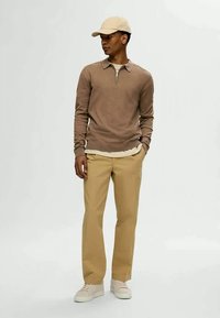 Man i beige keps, brun långärmad tröja med dragkedja i kragen, kaki byxor och beige sneakers, stående med ena handen i fickan.