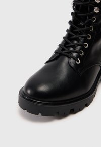 Botine negre din piele cu vârful rotund, suprafață texturată, ochiuri metalice și design cu șireturi. Sole din cauciuc masiv cu profil zimțat.