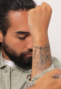 Juodas virvės apyrankė su mažuoju apvaliu mėlynu ir baltu karoliuku, dėvima ant riešo su matomais tatuiruotės raštais. Ranka laikoma priešais ranką.