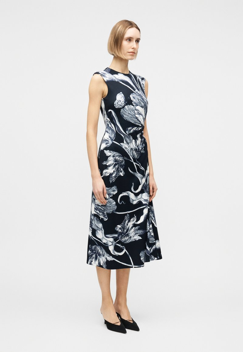 Woman standing in a sleeveless black midi dress with large white floral and butterfly prints, wearing black pointed-toe heels.
