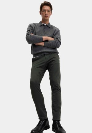 Grijze gebreide trui met een kraag, gecombineerd met donkergroene slim-fit broek en zwarte veterlaarzen. Model staat met gekruiste armen.