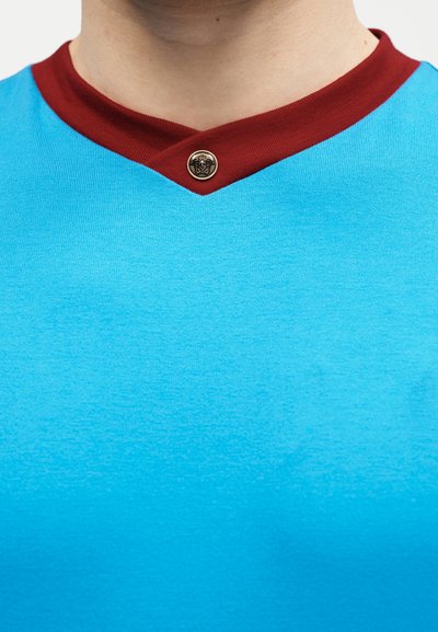 Gros plan sur une personne portant une chemise bleue avec un col en V bordeaux fixé par un bouton métallique rond orné d'un emblème de lion.