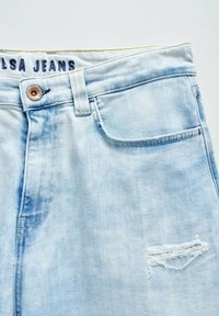 Vaqueros de mezclilla azul claro con un acabado desgastado, que presentan un bolsillo frontal, un desgastado roto en una pierna y una cinturilla con marca.