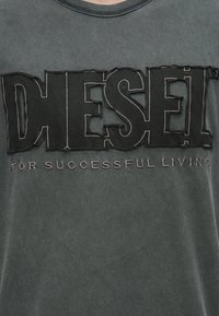 Fekete pamut póló, amelyen nagy betűkkel és varrással kidolgozott, struktúrált "DIESEL" logó található, alatta pedig az "A sikeres életért" felirat.