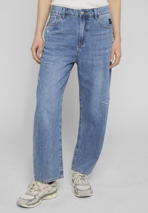 Personne portant un jean droit bleu clair délavé et des baskets blanches et grises, debout devant un fond uni.