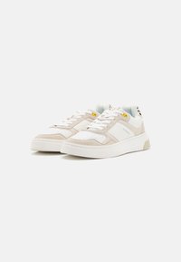 Light beige sneakers with white leather accents, textured suede details, round toe, flat white rubber sole, and yellow and blue hardware.