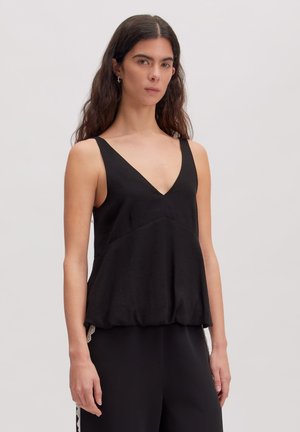 Vrouw met lang donker haar die een mouwloze zwarte top met V-hals en een zwarte broek draagt tegen een effen lichte achtergrond.