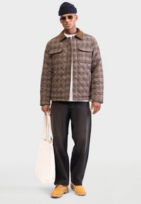 Mand iført ternet foret jakke, sort beanie, solbriller, sorte jeans, gule støvler, holdende en beige tote-bag, stående mod en hvid baggrund.