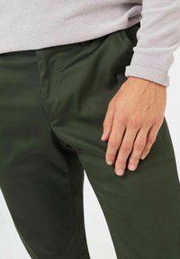 Mörkgröna byxor tillverkade av ett slätt, lätt material, med sidofickor och en platt frontdesign. Hand placerad på låret.