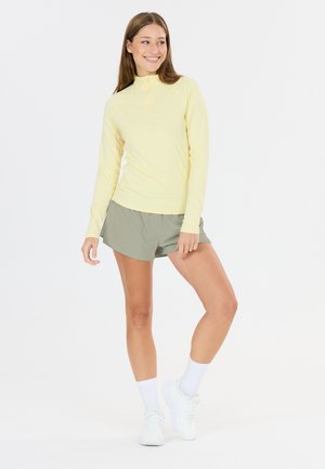 Junge Frau lächelt, trägt ein hellgelbes Langarmshirt, graue Shorts, weiße Socken und weiße Turnschuhe, steht vor weißem Hintergrund.