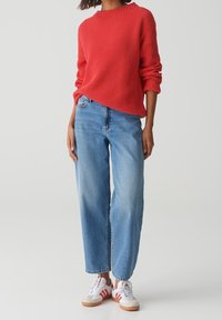Person, der einen roten Strickpullover, hoch taillierte hellblaue Jeans und weiße Sneaker mit roten Streifen trägt, steht vor einem einfarbigen Hintergrund.