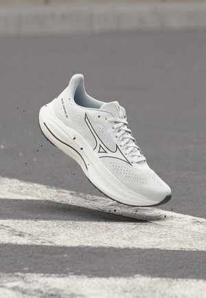 Chaussure de course Mizuno blanche en suspension au-dessus de l'asphalte, avec des graviers lâches soulevés, montrant la semelle et le logo latéral.