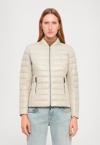 Padded zip-up jakna u svijetloj bež boji s kvadratnim dizajnom, stojećim ovratnikom i bočnim patentnim zatvaračima. Glatka tekstura, prilagođeni kroj.