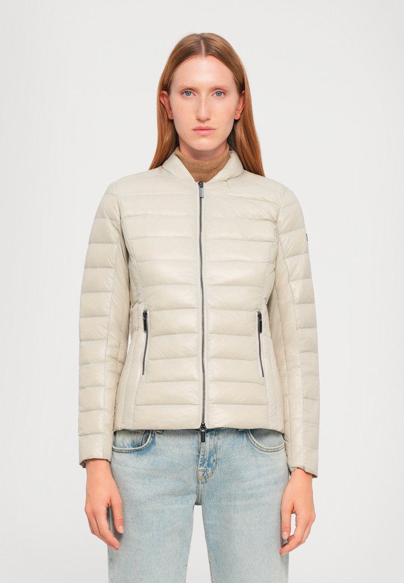 Padded zip-up jakna u svijetloj bež boji s kvadratnim dizajnom, stojećim ovratnikom i bočnim patentnim zatvaračima. Glatka tekstura, prilagođeni kroj.