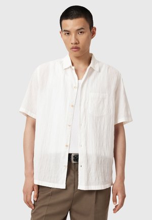 Camicia bianca a maniche corte realizzata in tessuto testurizzato, con chiusura a bottoni e una tasca sul petto, indossata sopra una canotta bianca.