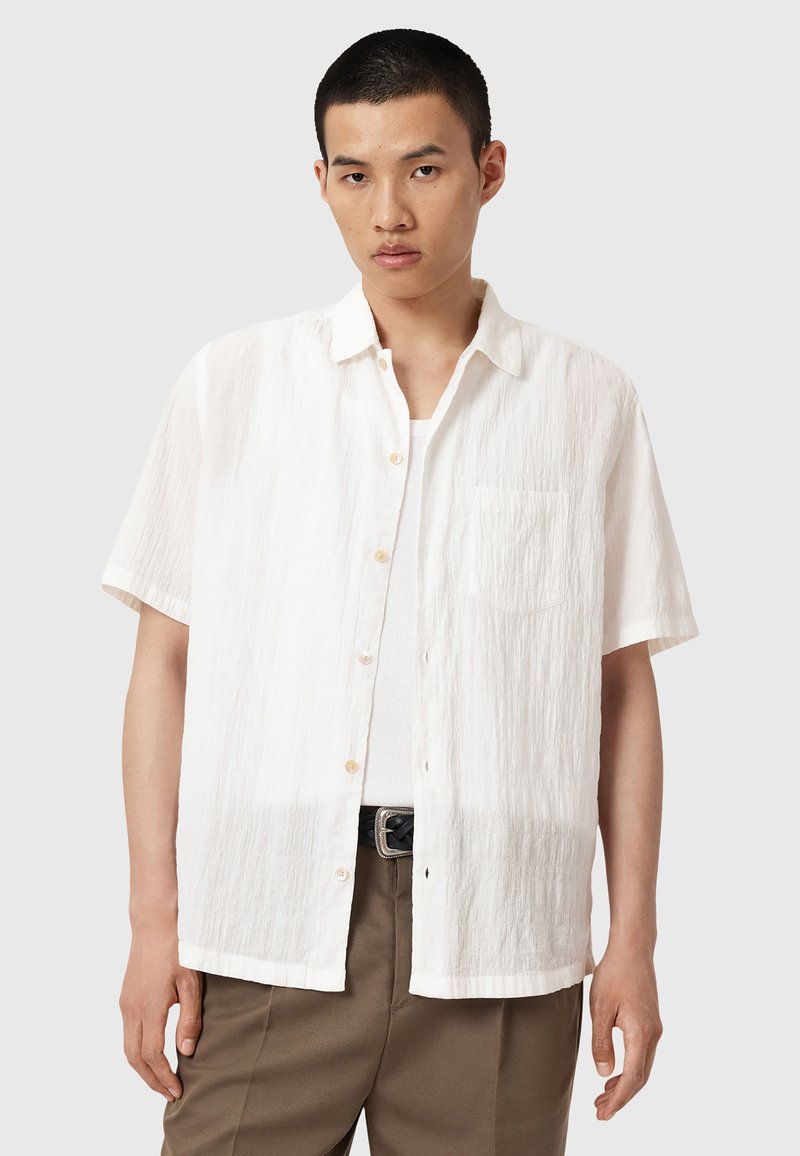 Chemise blanche à manches courtes en tissu texturé avec un front à boutons et une poche poitrine, portée sur un débardeur blanc.