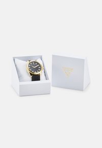Montre noire avec des accents dorés, présentée dans une boîte blanche avec un insert rembourré. La boîte arbore un logo doré sur le devant.