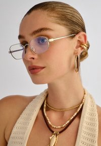 Γυναίκα με μαλλιά χτενισμένα προς τα πίσω φοράει γυαλιά με μπλε απόχρωση και ορθογώνιο σχήμα, χρυσούς κρίκους σκουλαρίκια, στρωμένα κολιέ από χάντρες και κορδόνια, καθώς και ένα κρεμ πλεκτό τοπ.