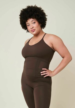 Donna con capelli ricci che indossa un top marrone attillato e leggings, in piedi con una mano sul fianco su uno sfondo semplice.