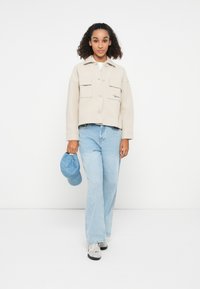 Beige cropped jacket with button closure and two front pockets. Light blue wide-leg jeans. Holding a light blue cap; grey sneakers complete the look.