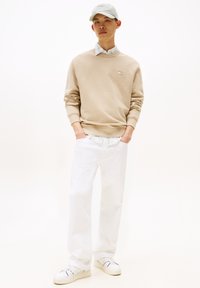 Sudadera beige con cuello redondo, pantalones vaqueros holgados blancos y gorra verde pálido. Zapatillas blancas con acentos azules completan el atuendo.